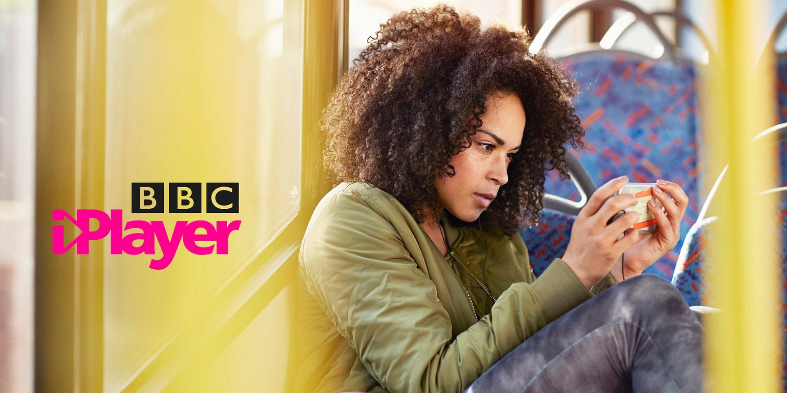 A woman with curly hair and a green jacket is sitting on a bus, holding a cup of coffee and looking at her phone.