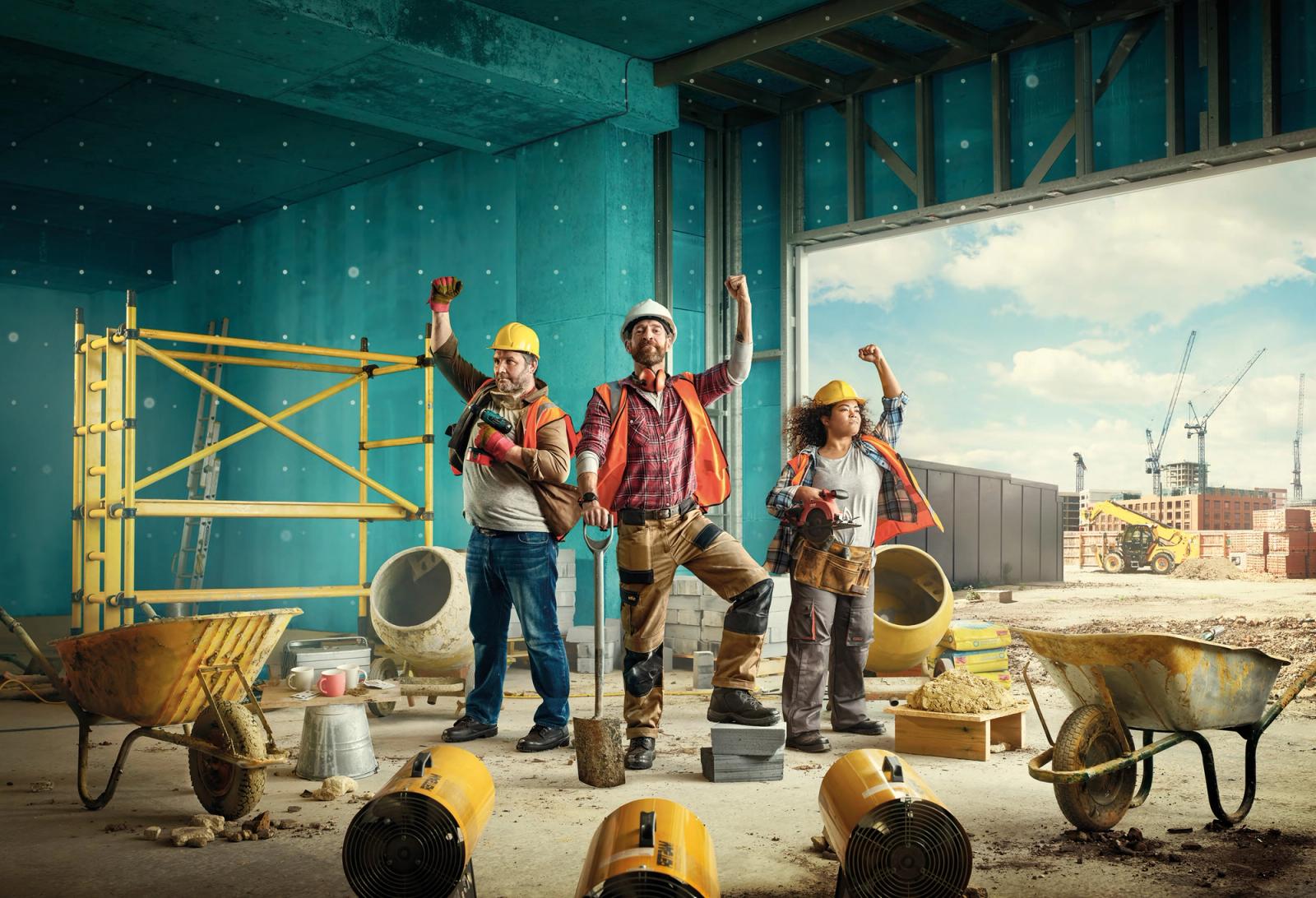 A group of workers stand in front of a blue-painted wall with a large window, wearing hard hats and high-visibility vests, surrounded by construction equipment.