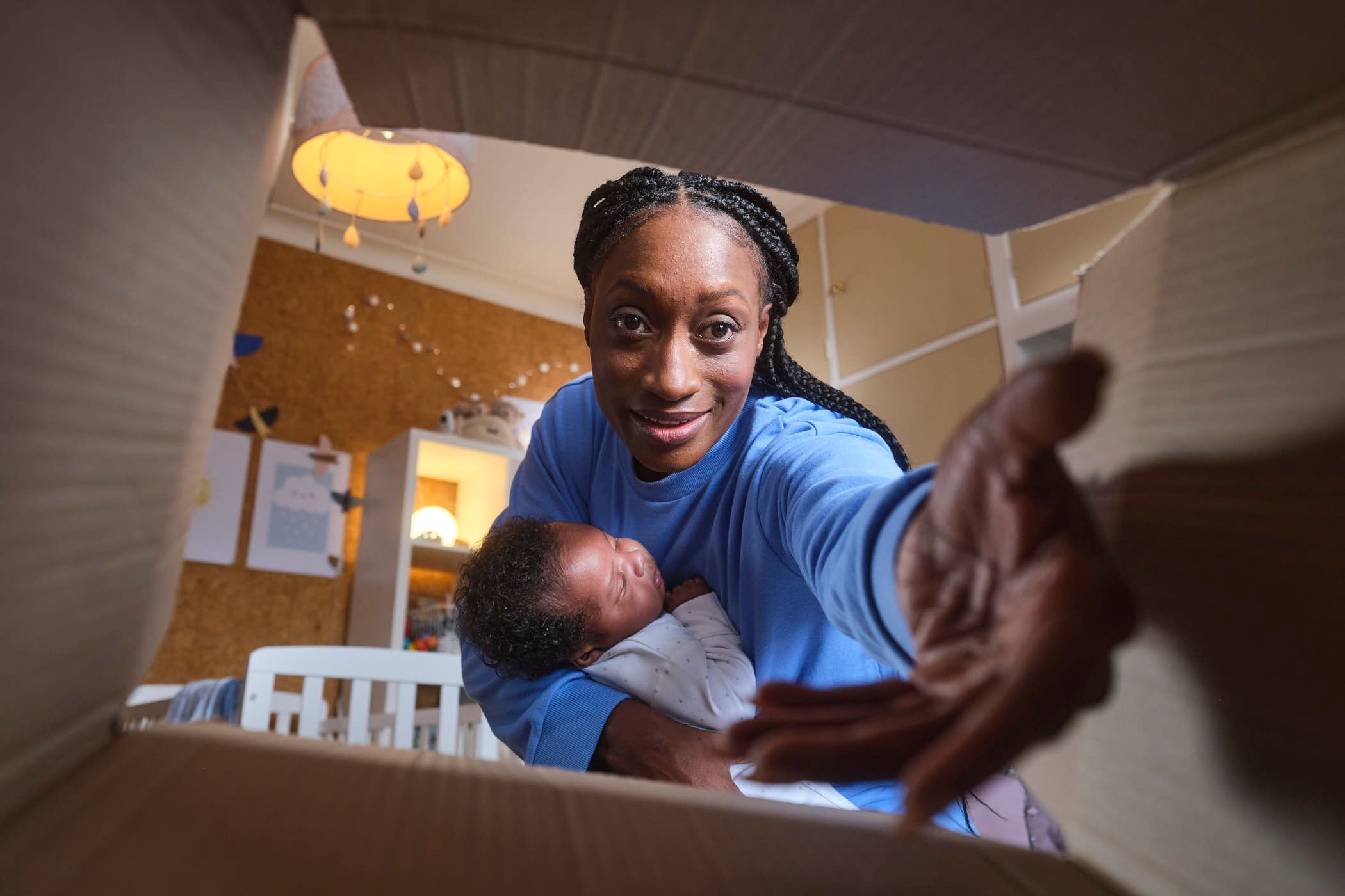 A woman with dark skin and braided hair holds a baby in her arms, smiling at the camera through an open cardboard box. The room behind her is a nursery with a white crib, a.