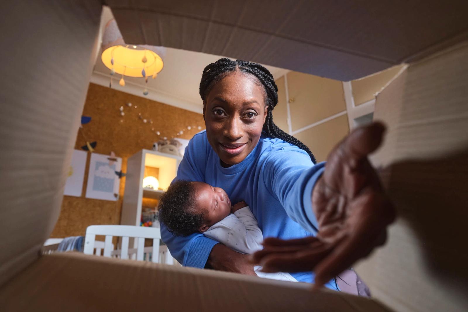 A woman with dark skin and braided hair holds a baby in her arms, smiling at the camera through an open cardboard box. The room behind her is a nursery with a white crib, a.