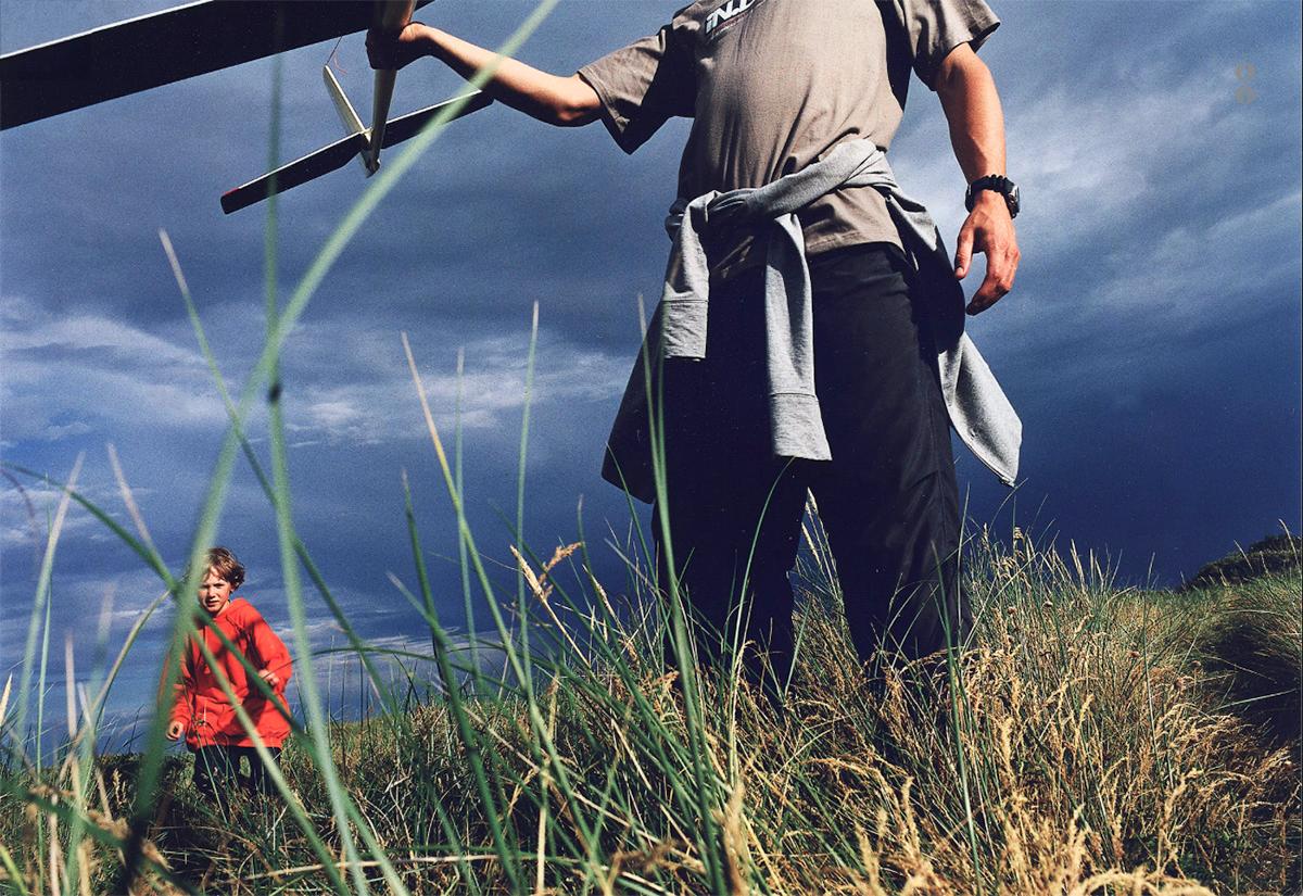 A man in a grey t-shirt and black trousers is holding a toy airplane in a field of tall grass, with a child in a red jacket in the background.