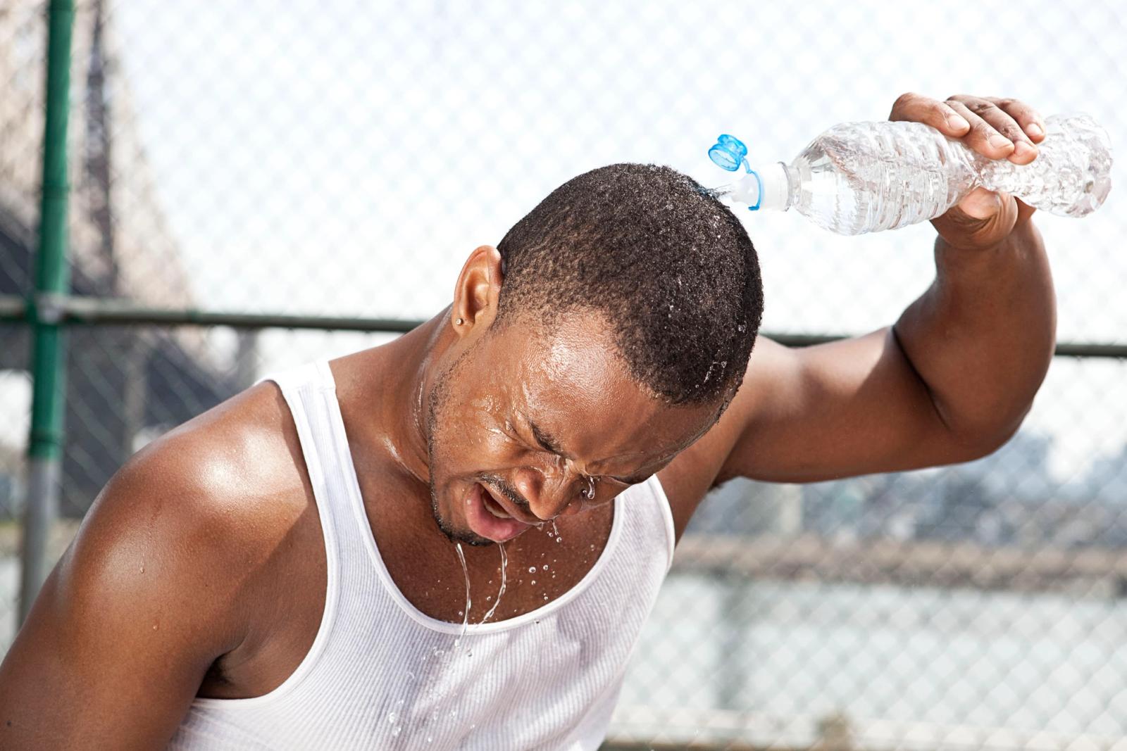 A man in a white tank top is pouring water from a plastic bottle over his head, with water droplets falling from his face and a chain-link fence in the background.