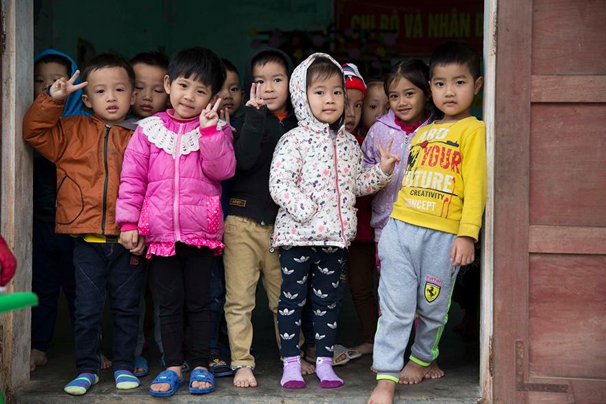 A group of young children standing in a doorway, wearing casual clothing and making peace signs with their hands.