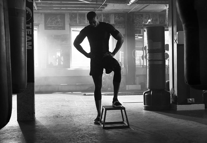 A man in a dark t-shirt and shorts is standing on a small step stool in a dimly lit gym, with a large window in the background.