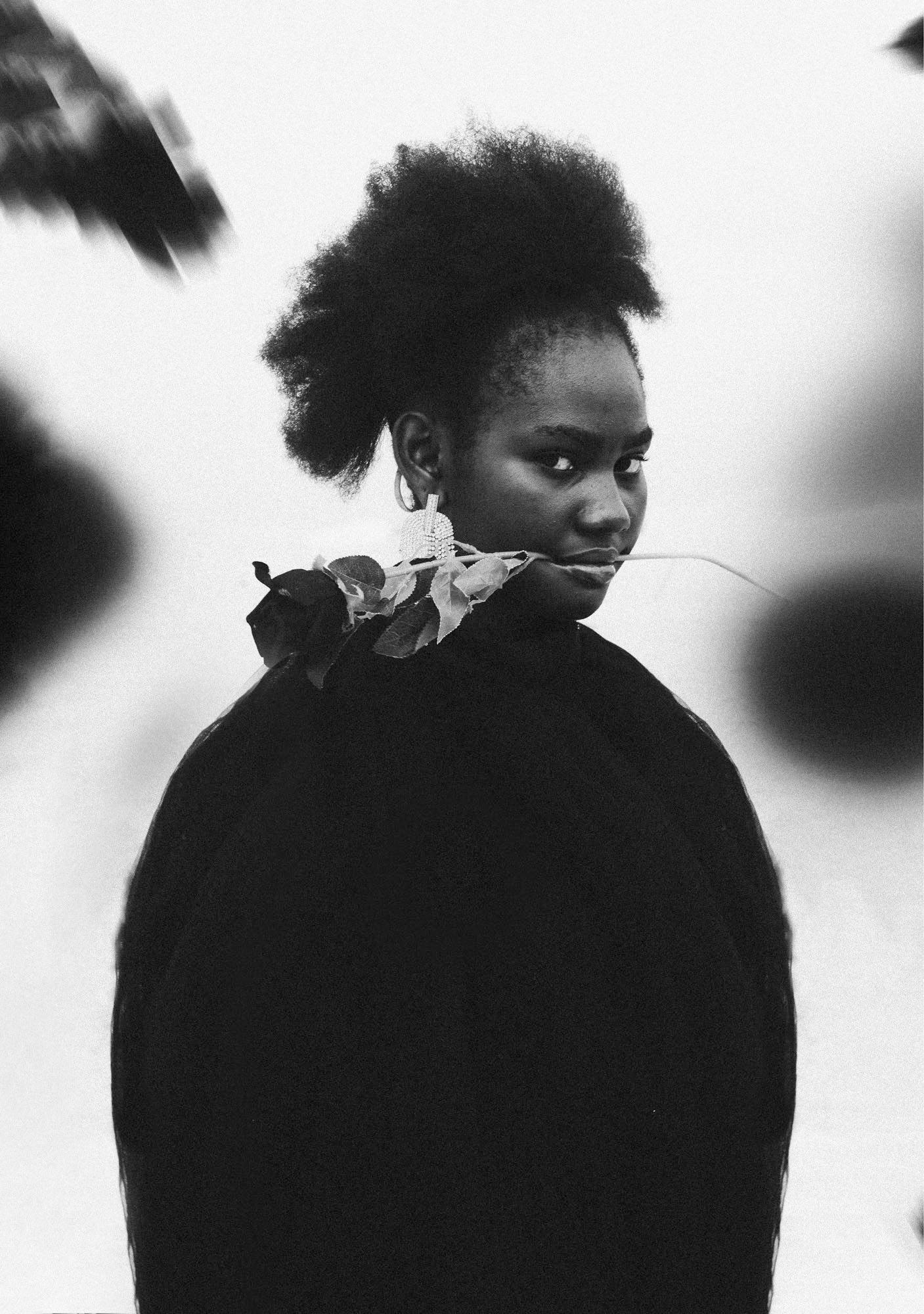 A young woman with a natural hairstyle and a dark, simple dress, holding a flower in her mouth.
