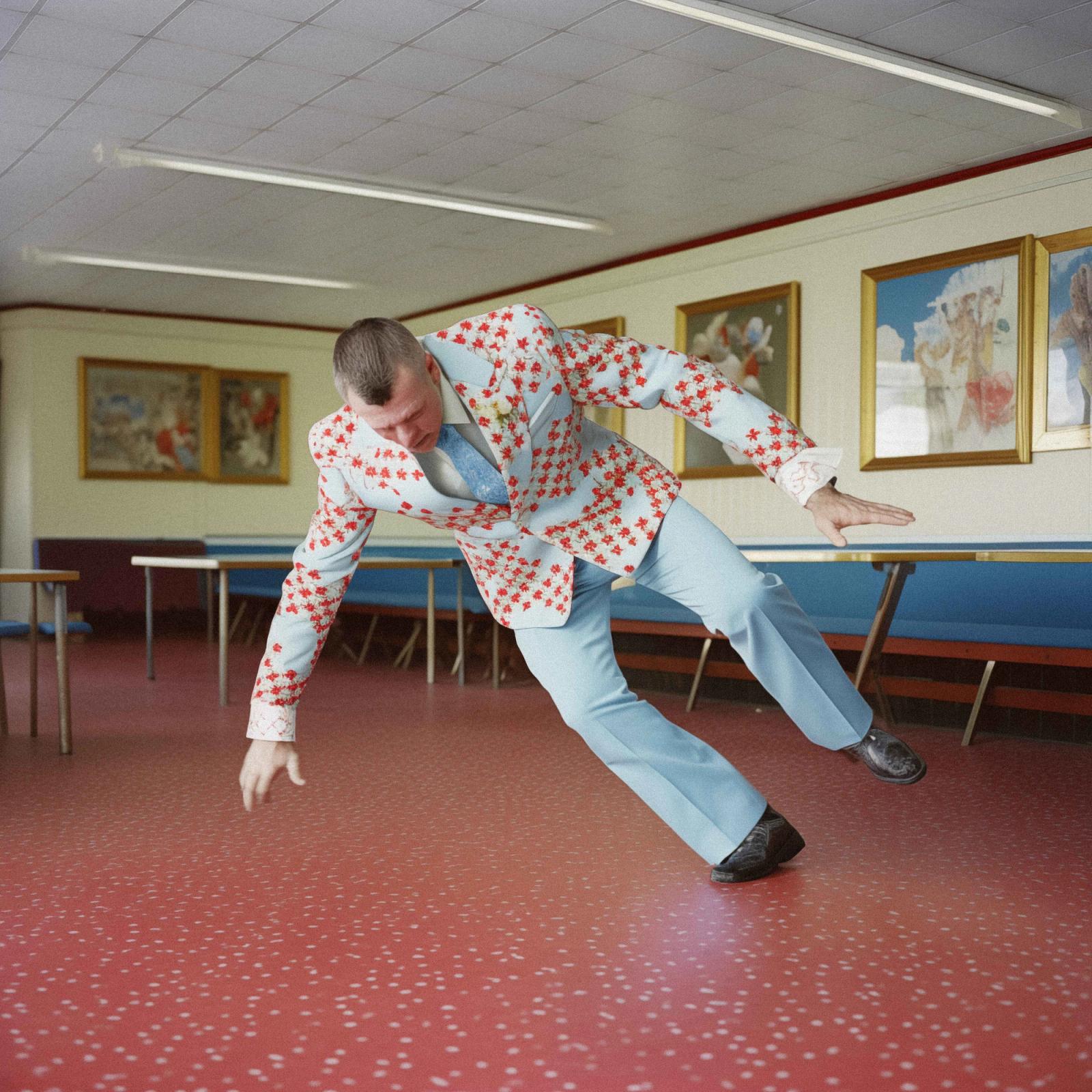 A man in a white suit with red flowers, wearing a blue tie, is dancing in a room with a red floor and blue benches.