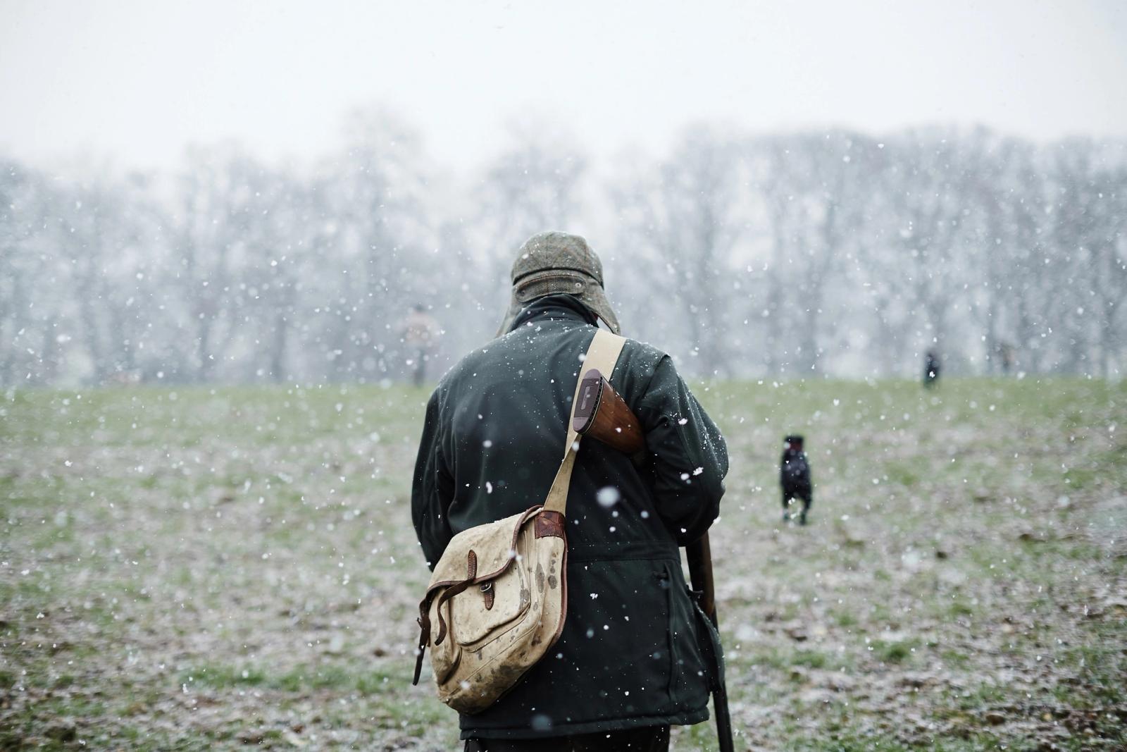 A man in a black coat and hat, carrying a rifle, walking through a snowy field with a dog in the distance.