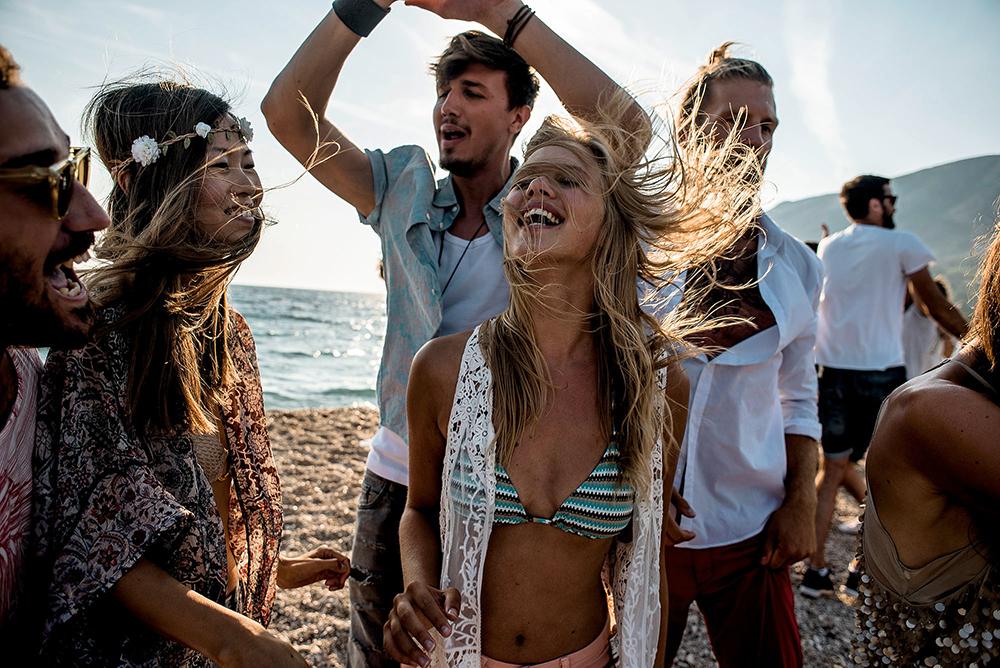 A group of young adults, likely tourists, are dancing and having fun on a beach in Croatia, with the sun setting in the background and the sea in the distance.