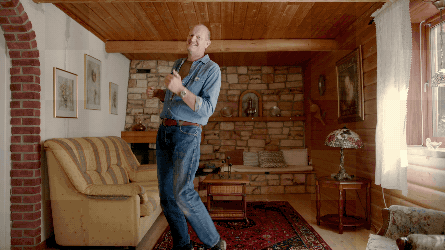 A man in a blue denim shirt and jeans standing in a living room with a couch, a coffee table, and a fireplace.
