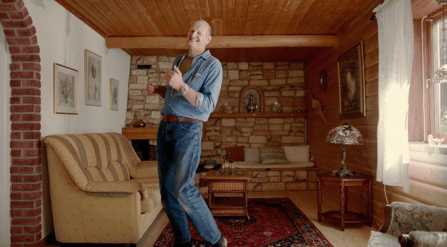 A man in a blue denim shirt and jeans standing in a living room with a couch, a coffee table, and a fireplace.