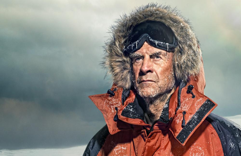 A man in a thick, orange and black parka with a fur-lined hood, snow-covered and looking out into the distance.