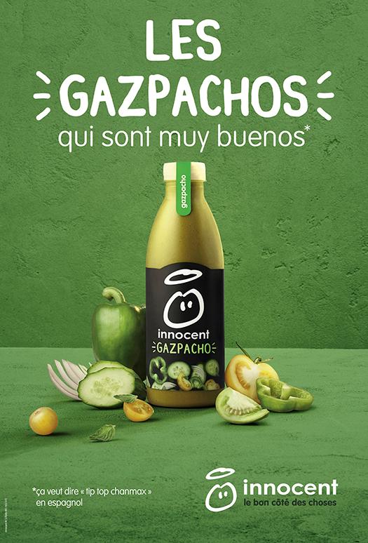 A bottle of gazpacho with various vegetables and fruits surrounding it, including a cucumber, a tomato, and a green apple.