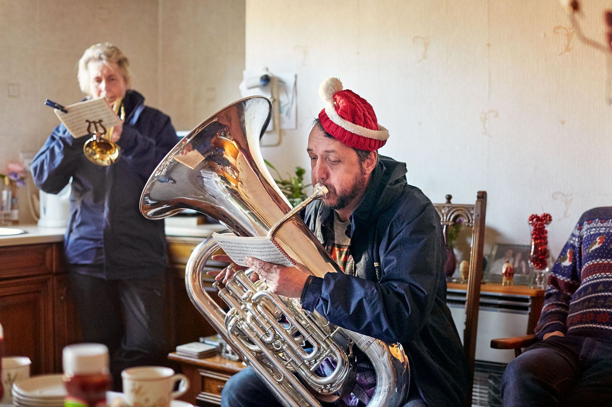 A man in a red Santa hat and a blue jacket playing a tuba in a living room, with a woman in the background also playing a trumpet.