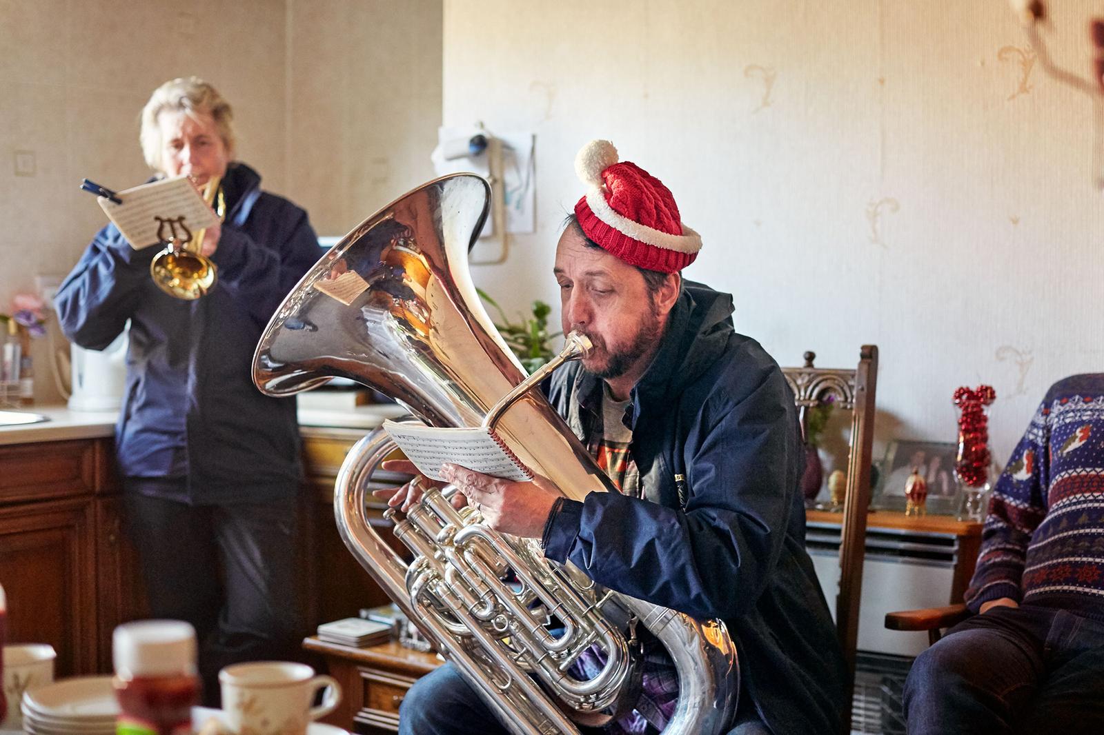 A man in a red Santa hat and a blue jacket playing a tuba in a living room, with a woman in the background also playing a trumpet.