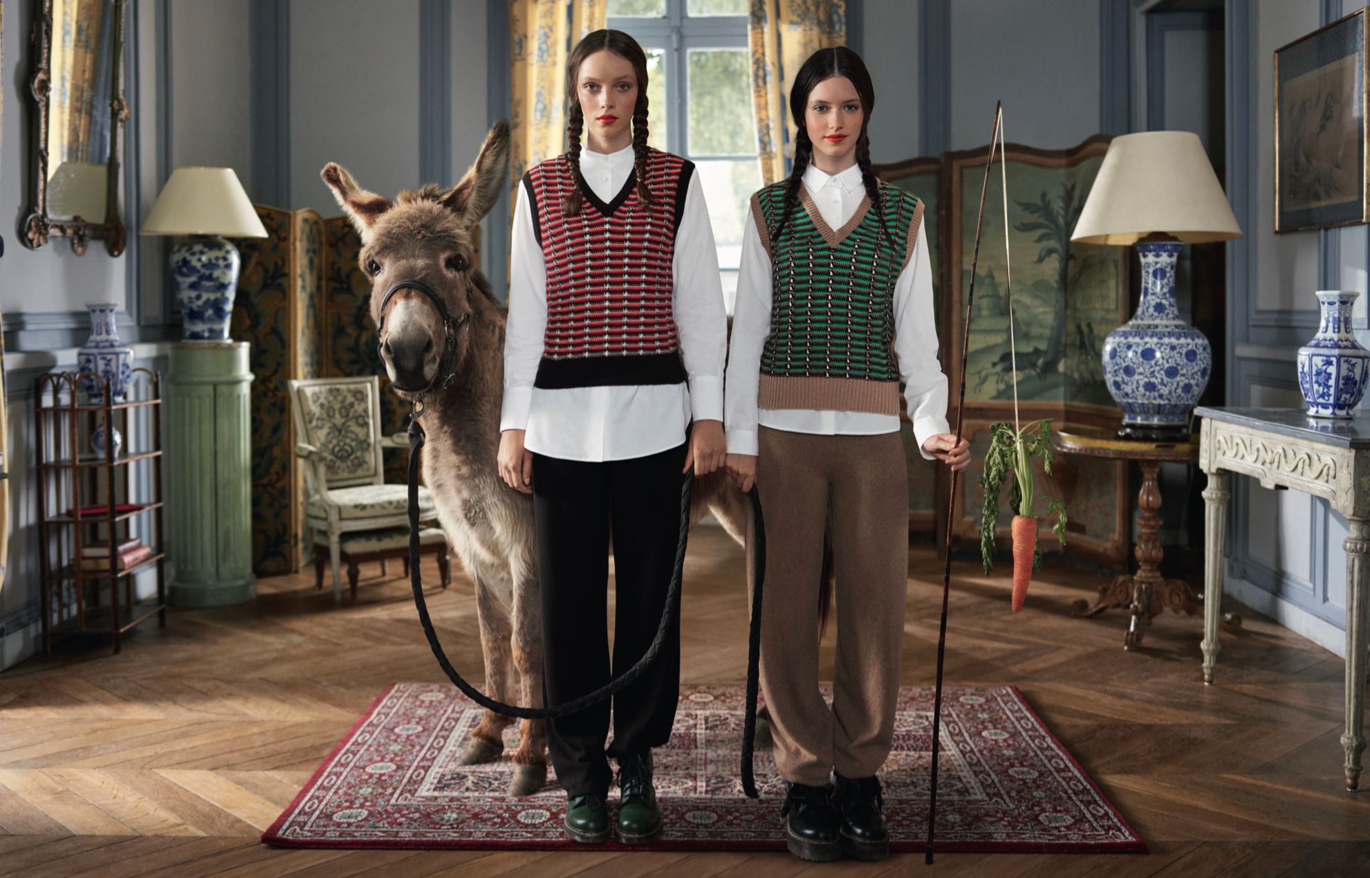 Two women in period clothing hold a long pole with a carrot, standing with a donkey in a room.