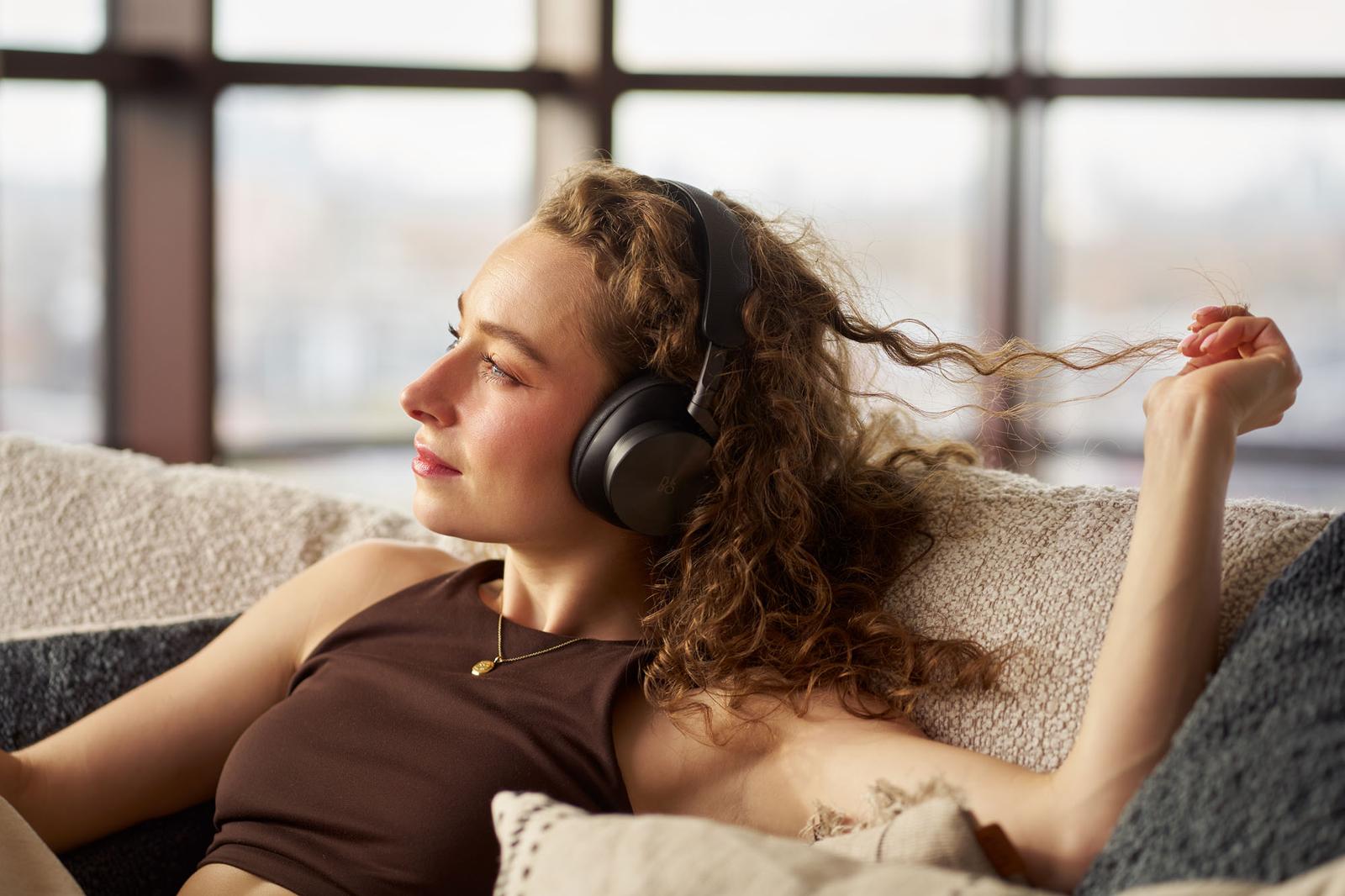 A woman sits on a couch wearing large black headphones and appears relaxed.