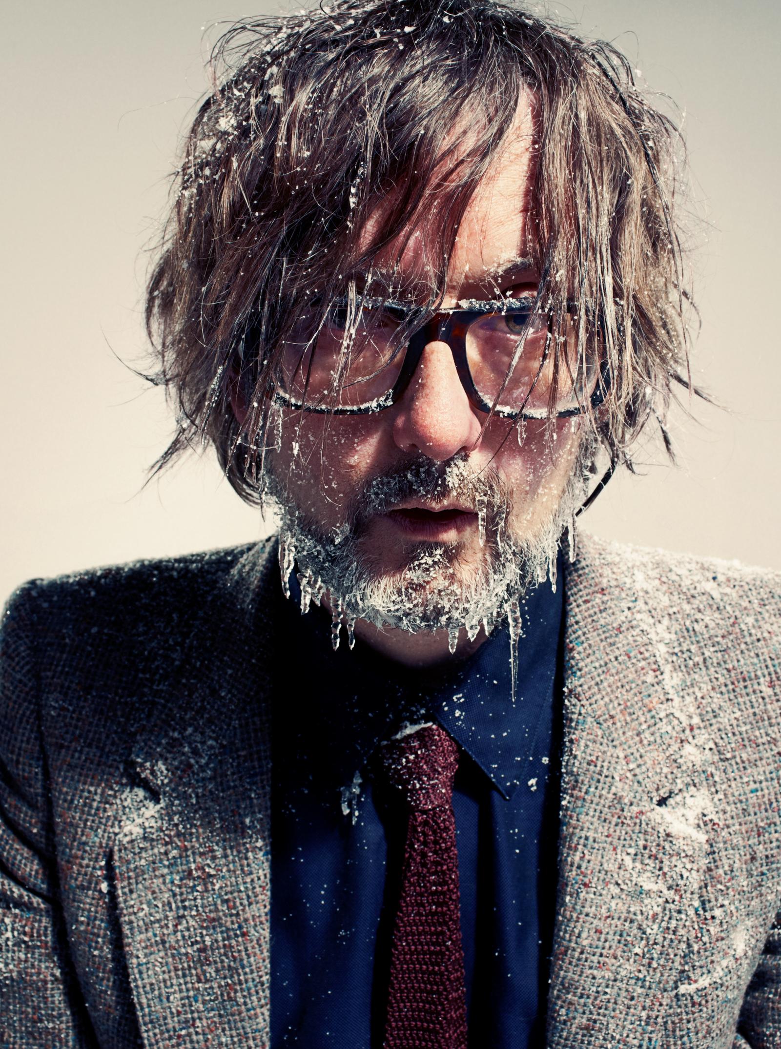 A man with long, wet, and icy hair and beard, wearing a tweed jacket, blue shirt, and red tie, with ice and snow covering his face and hair.