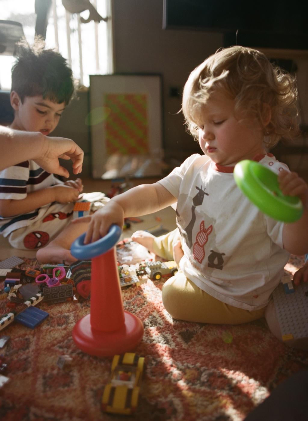 Two young children playing with a variety of toys on a rug, including a red cone, a blue ring, and a yellow car.