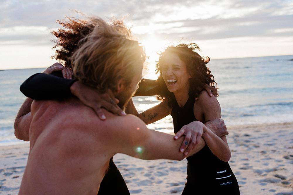 A group of people, including a woman with curly hair and a man with a beard, are embracing and laughing on a beach at sunset.