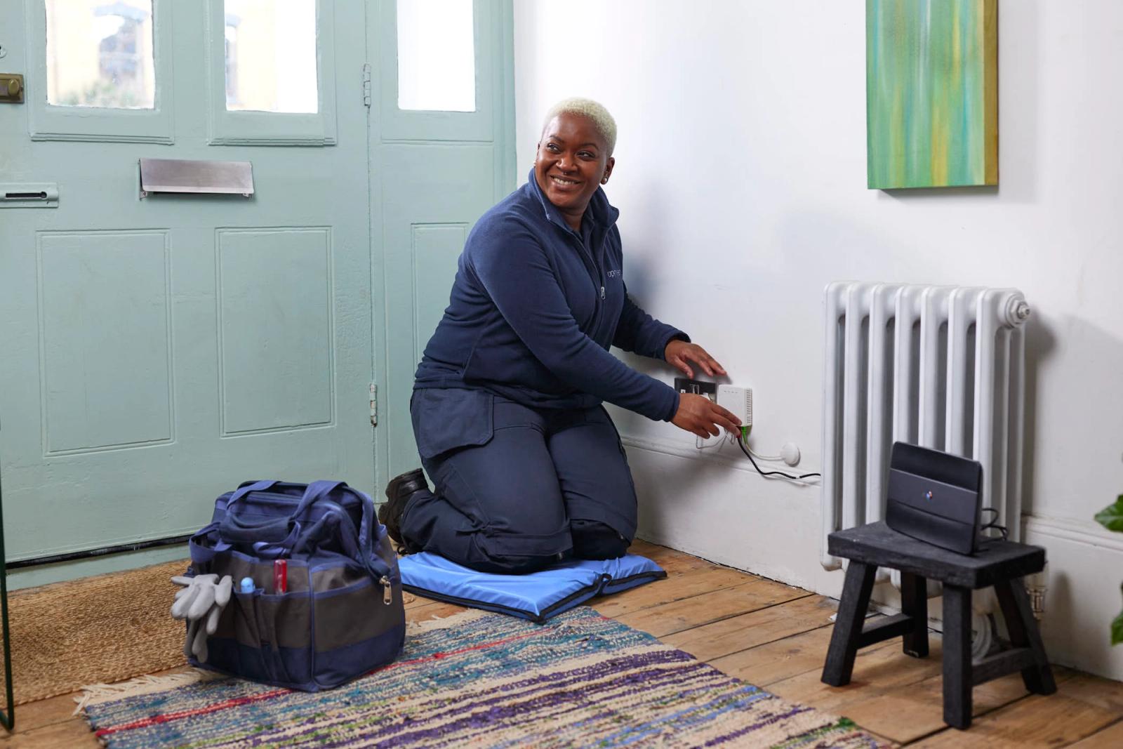 A woman with short blonde hair and a navy blue uniform kneels on a blue cushion in front of a white wall. She is smiling and pointing at a white device on the wall with her left.