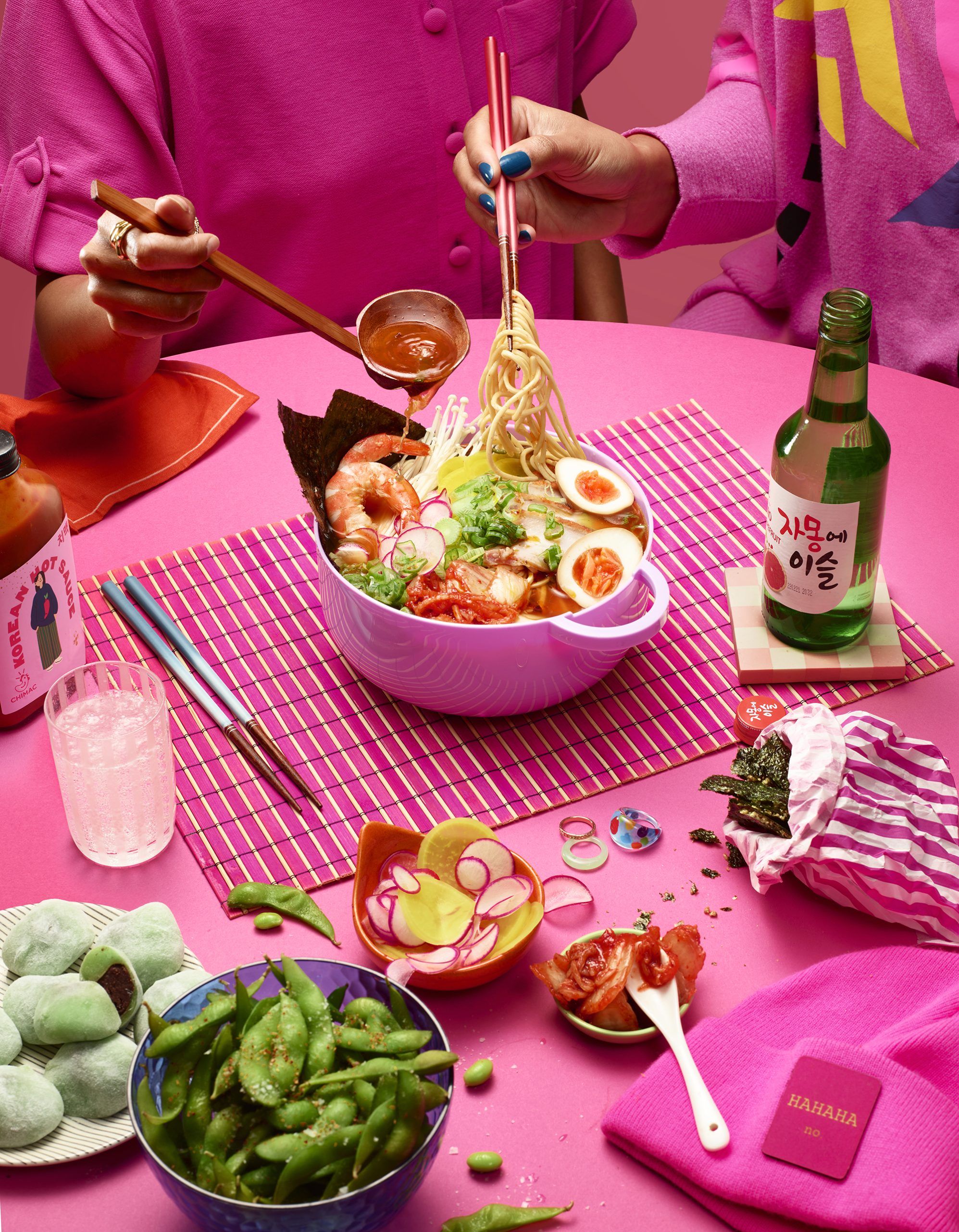 A woman in a pink shirt is lifting a noodle from a bowl of ramen. She has blue-painted nails and is using chopsticks to lift the noodle, which is covered in broth and various.