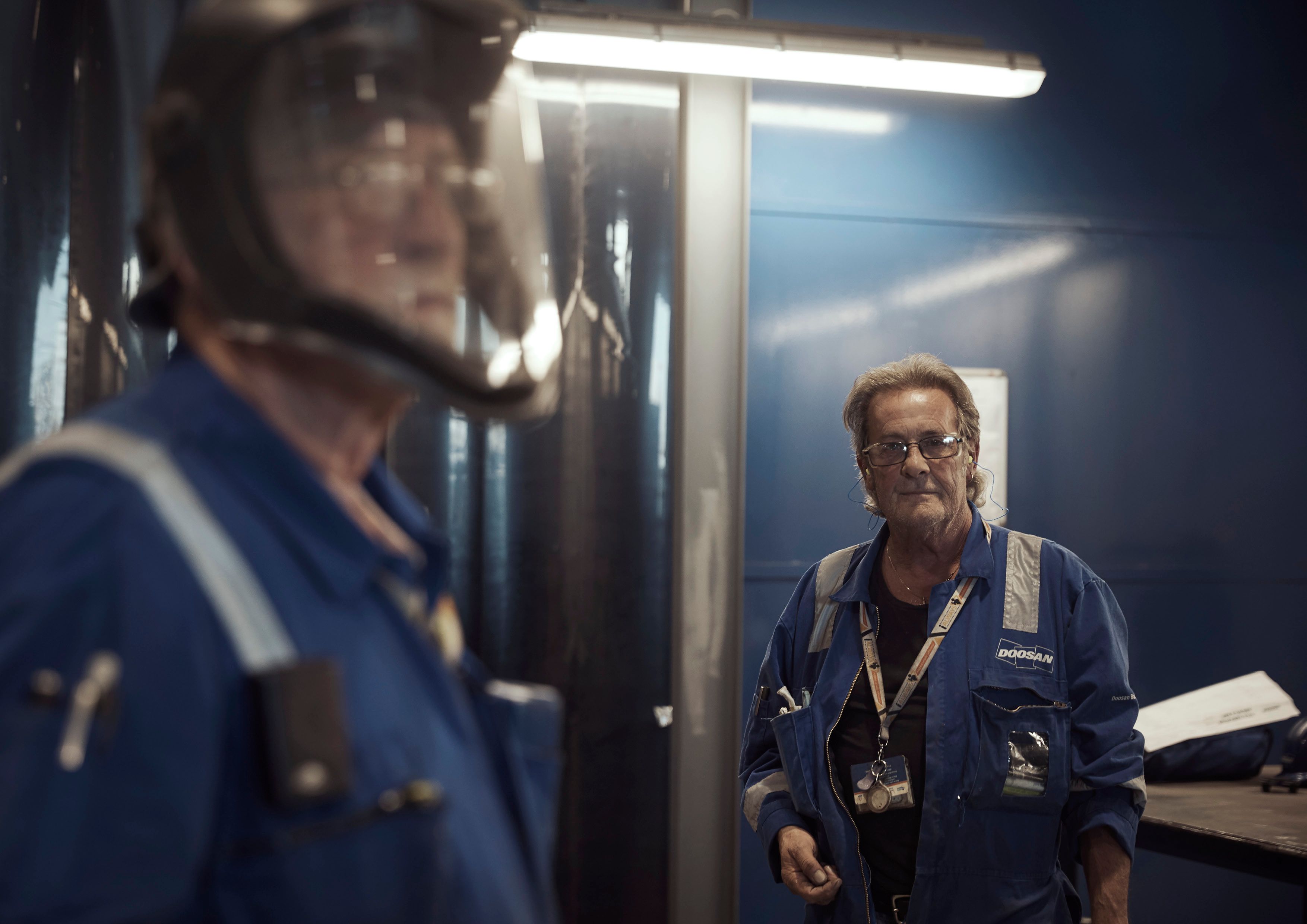 Two men in blue uniforms, one with a helmet, in an industrial setting.