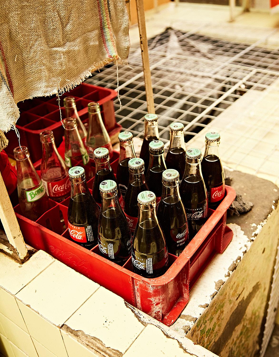 A red plastic crate holds 18 glass bottles of varying sizes and shapes on a white-tiled surface.