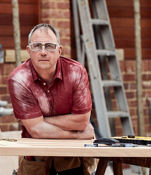 A man in a red shirt with white dust or powder on his clothes, standing in a workshop or construction site, with a ladder and tools in the background.
