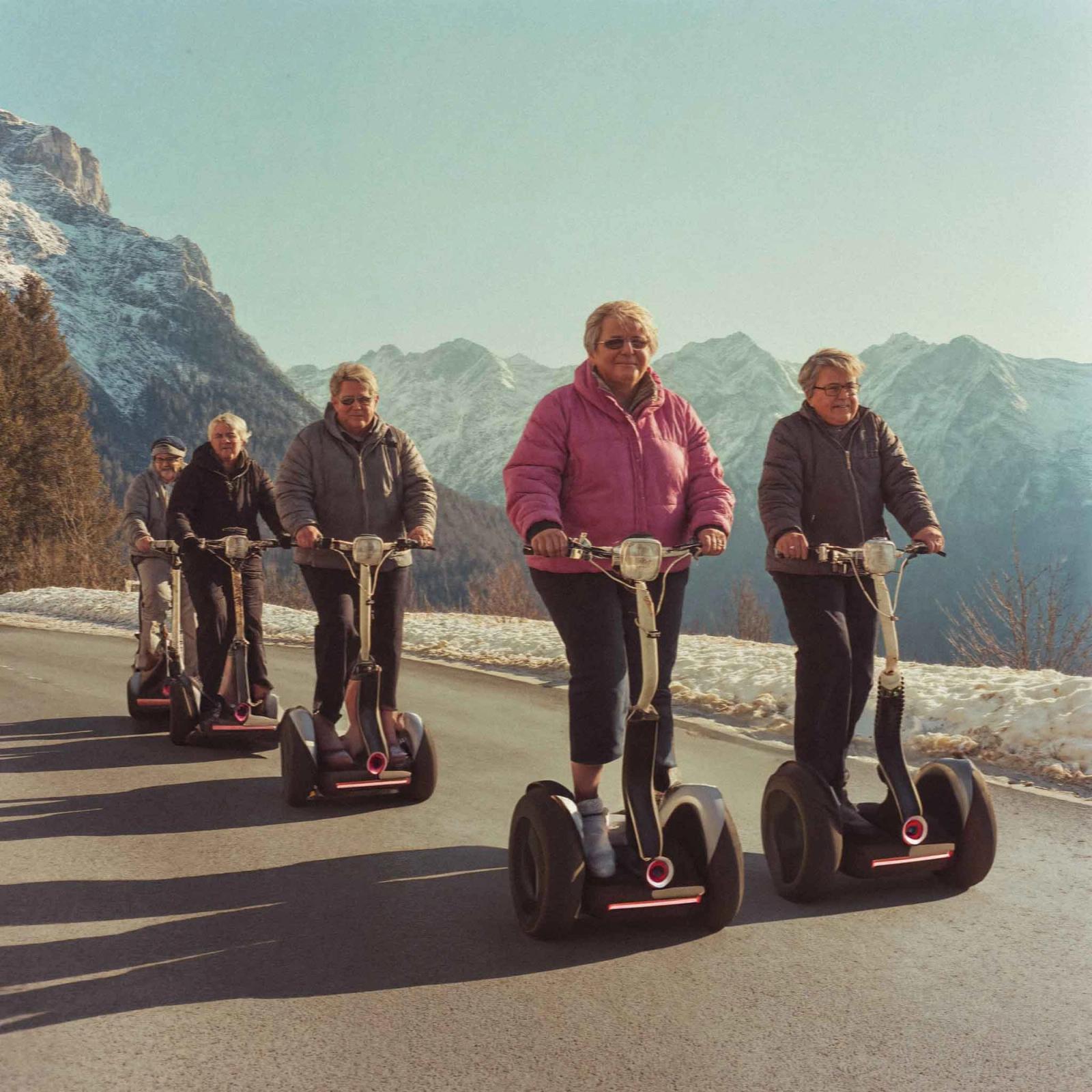 A group of older adults, likely tourists, wearing warm clothing and riding Segway-like vehicles, known as Segways, on a paved road in a mountainous, snow-covered landscape.