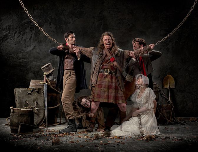 A group of actors, including a woman in a white wedding dress and a man in a kilt, are chained and being pulled by chains in a dark, dungeon-like setting.