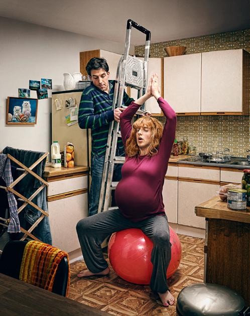 A woman in a red shirt and grey pants is sitting on a red exercise ball in a kitchen, with a man standing behind her holding a ladder.