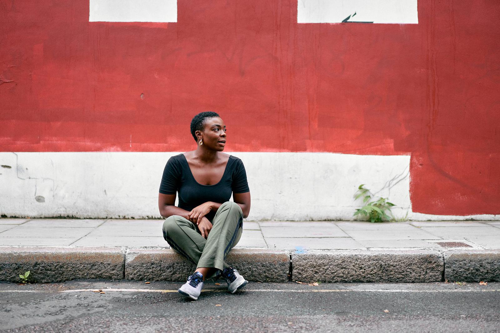 A woman with short hair and a black top sits on a curb, looking to the side.