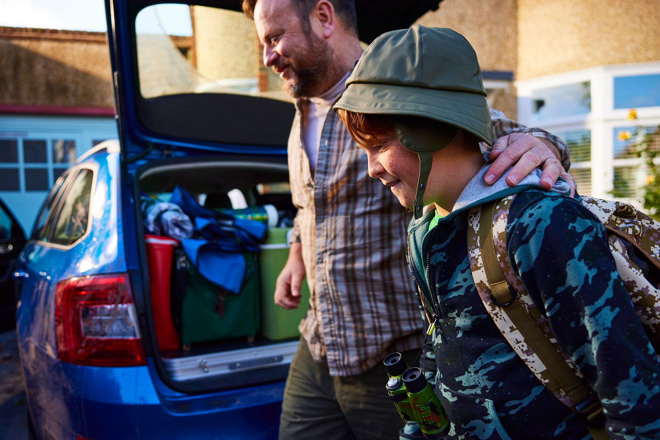 A man and a boy are standing in front of a blue car with its trunk open, packed with luggage. The man is wearing a plaid shirt and has his arm around the boy's shoulders. The boy.