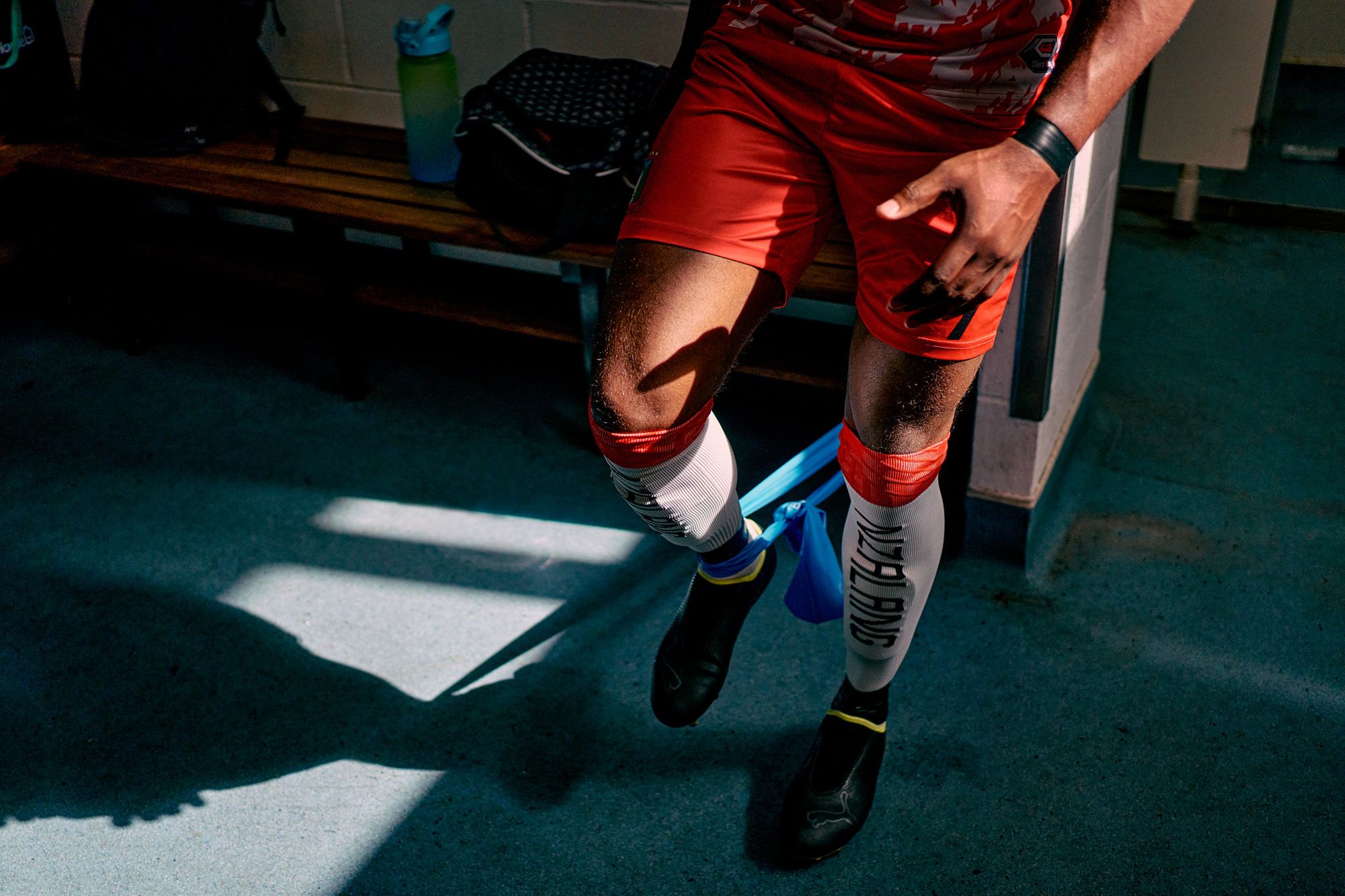 A person in a football jersey and football boots is sitting on a bench in a locker room, with a water bottle and a bag in the background.