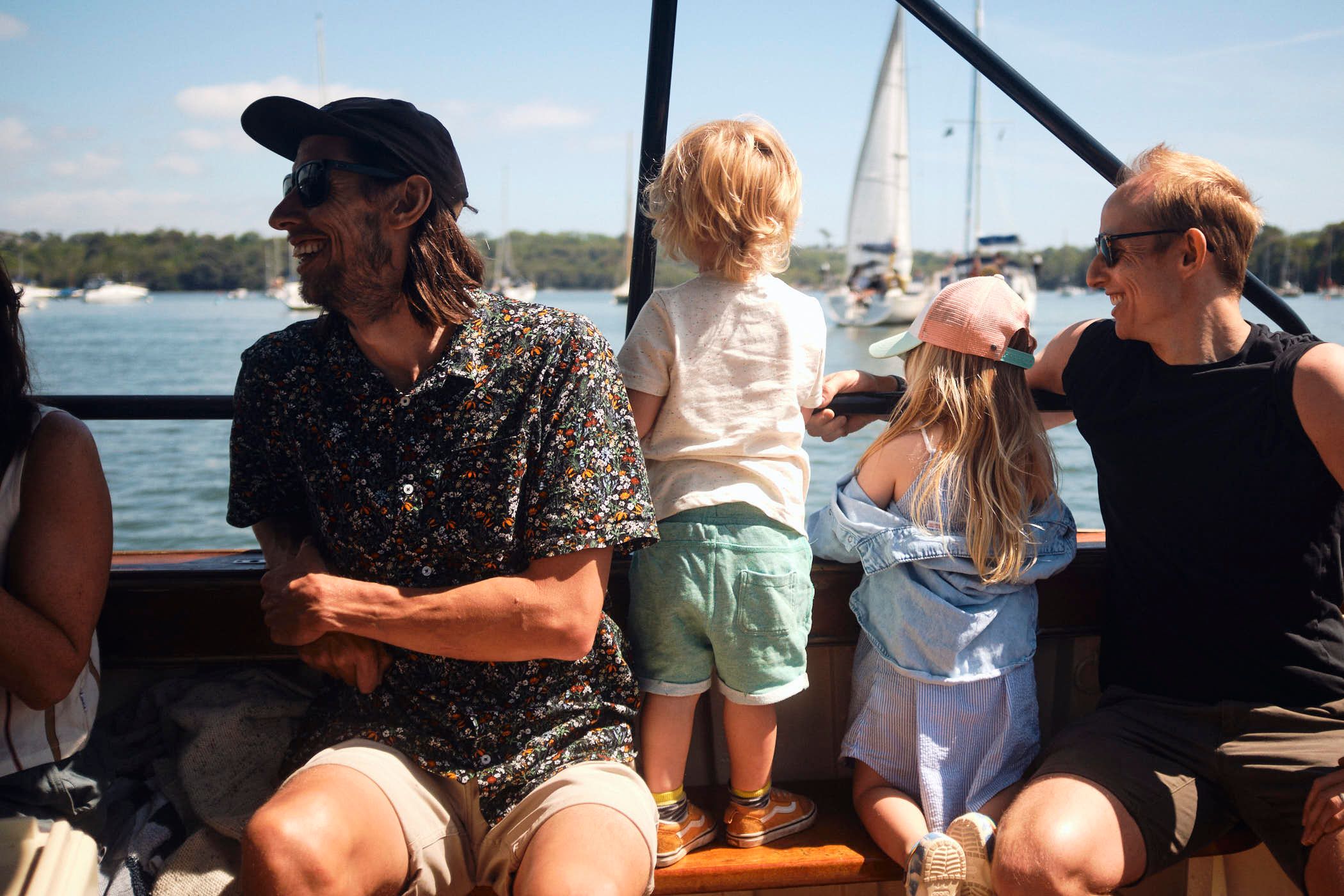 A man and a woman, both wearing casual summer clothing, sit on a boat with two young children. The man, with a beard and long hair, wears a black cap and a floral shirt. The.