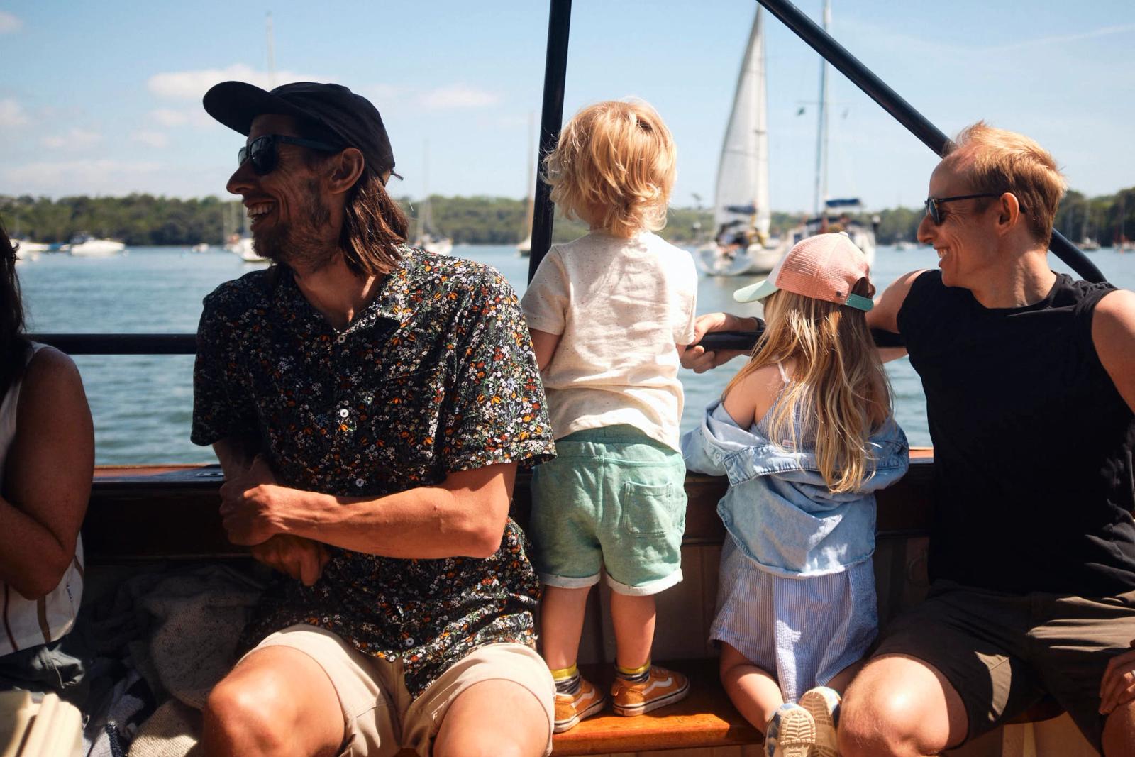 A man and a woman, both wearing casual summer clothing, sit on a boat with two young children. The man, with a beard and long hair, wears a black cap and a floral shirt. The.