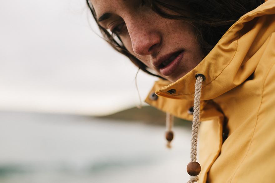 A woman wearing a yellow raincoat, standing in front of a blurred ocean background.