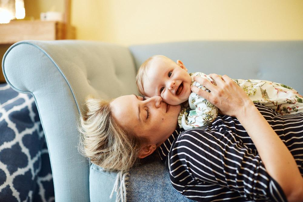 A woman with blonde hair and a black-and-white striped shirt is lying on a couch, holding a baby on her chest. The woman and the baby are both smiling and looking at the camera.