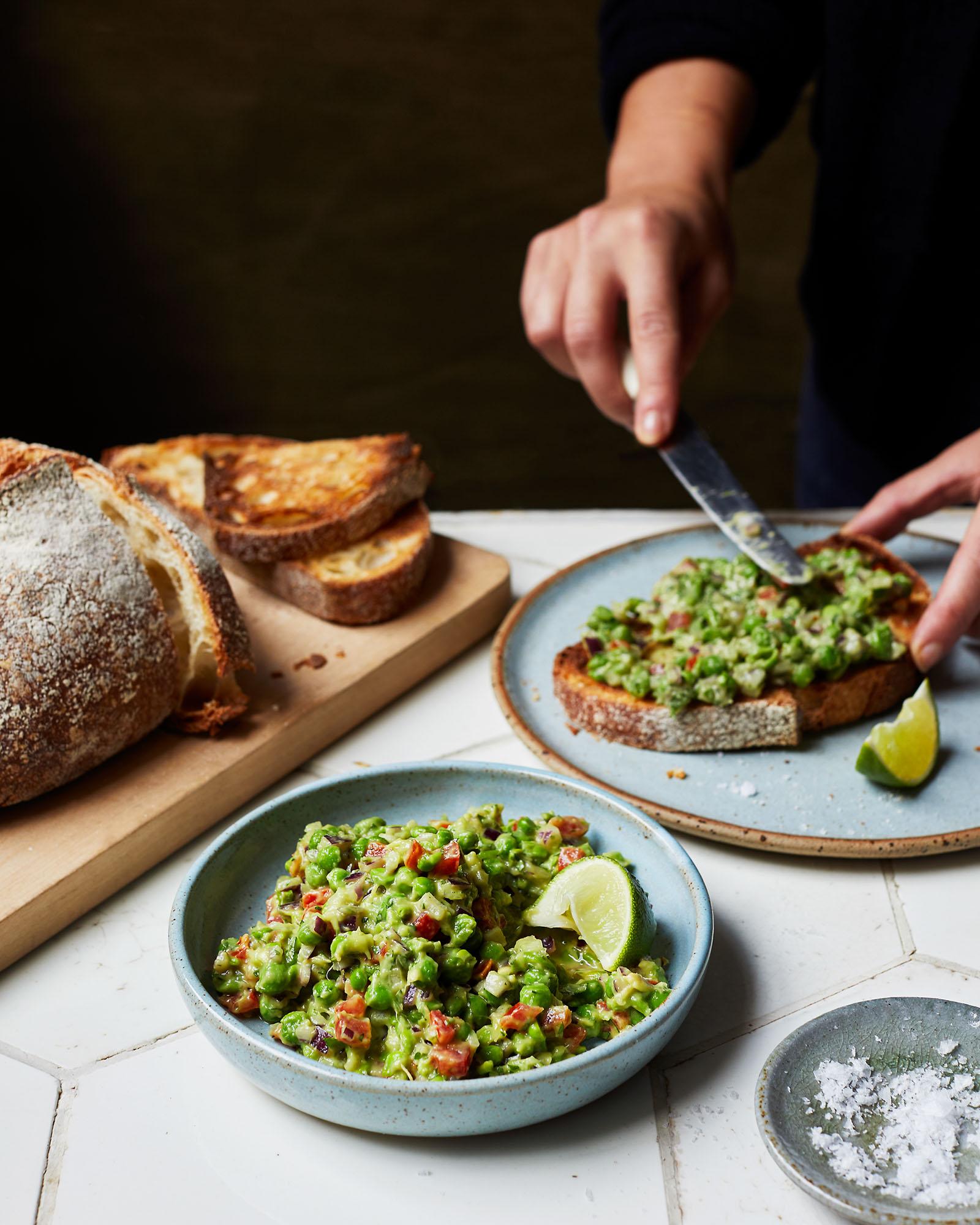 A person prepares guacamole with peas and avocado, focusing on the ingredients and hands.