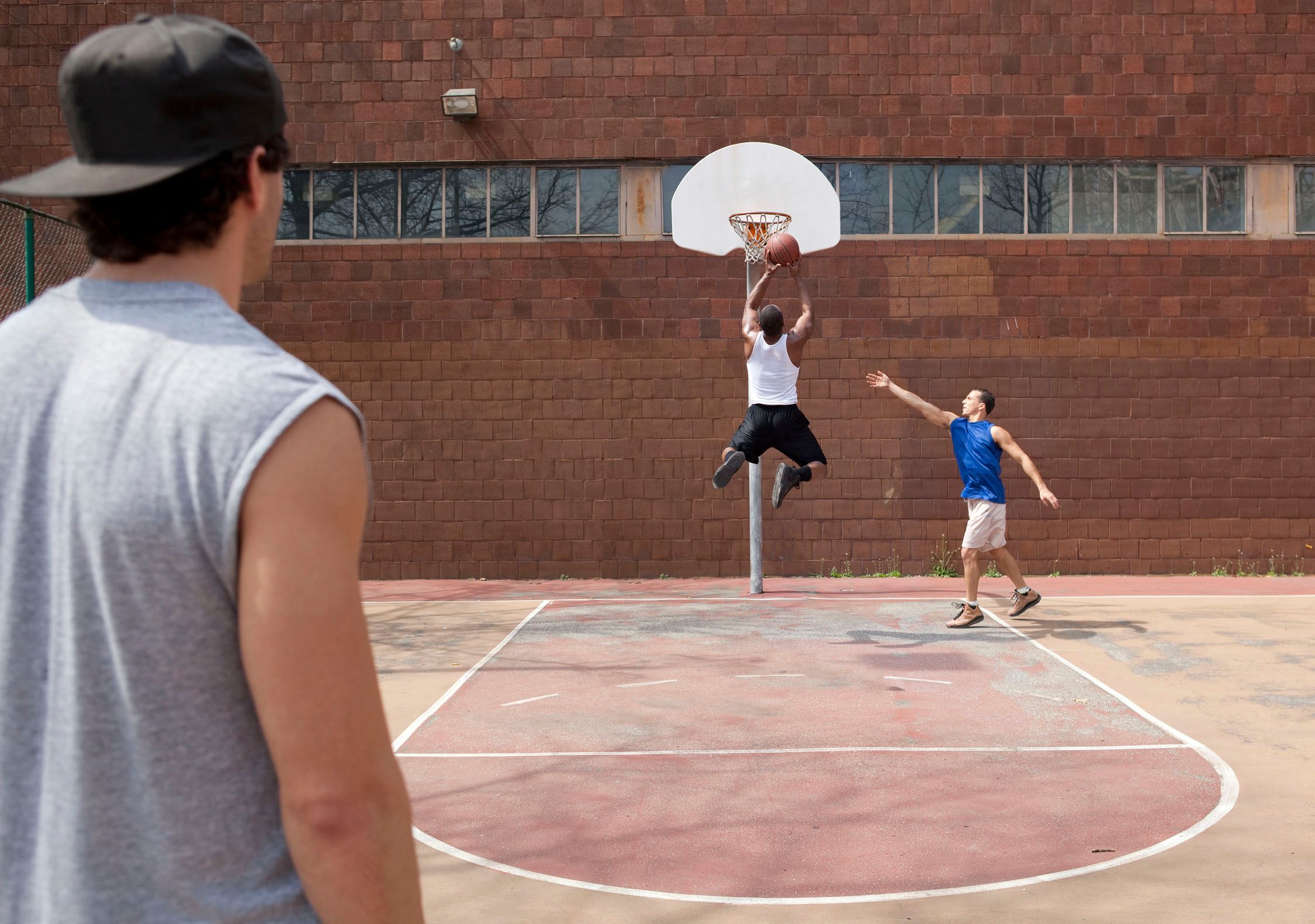 A man watching two players play basketball outdoors.