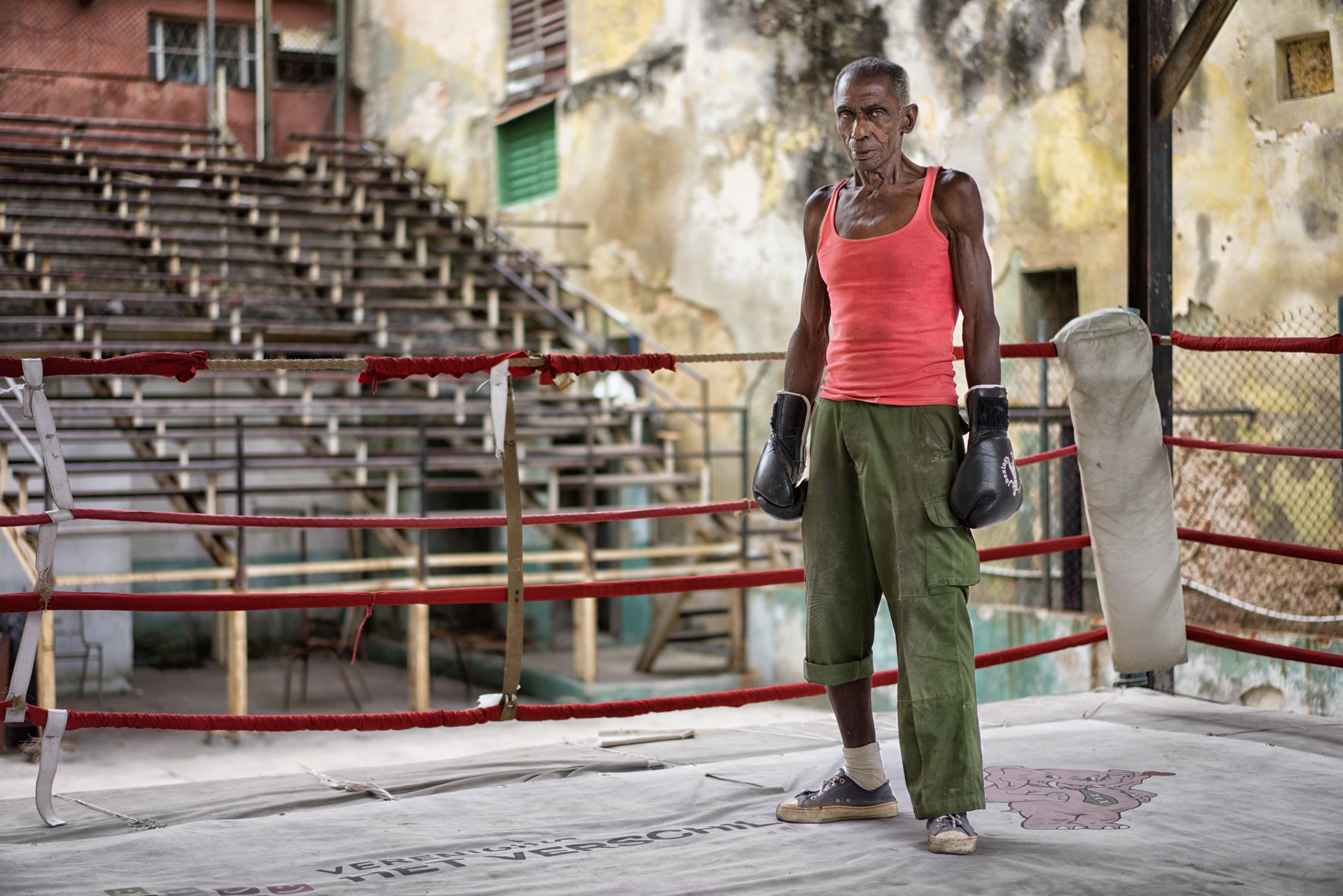 A man in a red tank top, green cargo pants, and boxing gloves stands in an empty, dilapidated boxing ring.