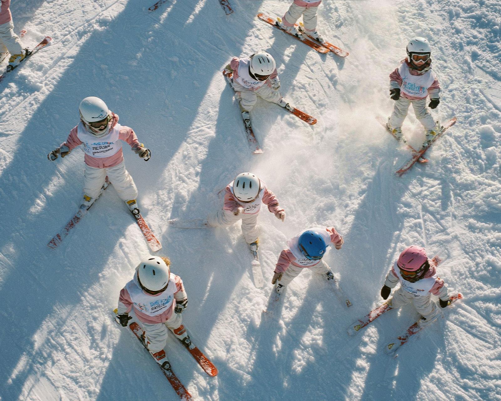 A group of young children in matching pink and white ski suits, wearing helmets and holding skis, standing on a snowy slope.