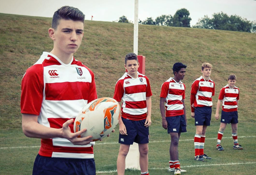 A group of young boys in red and white striped rugby jerseys standing on a rugby field, holding a rugby ball.