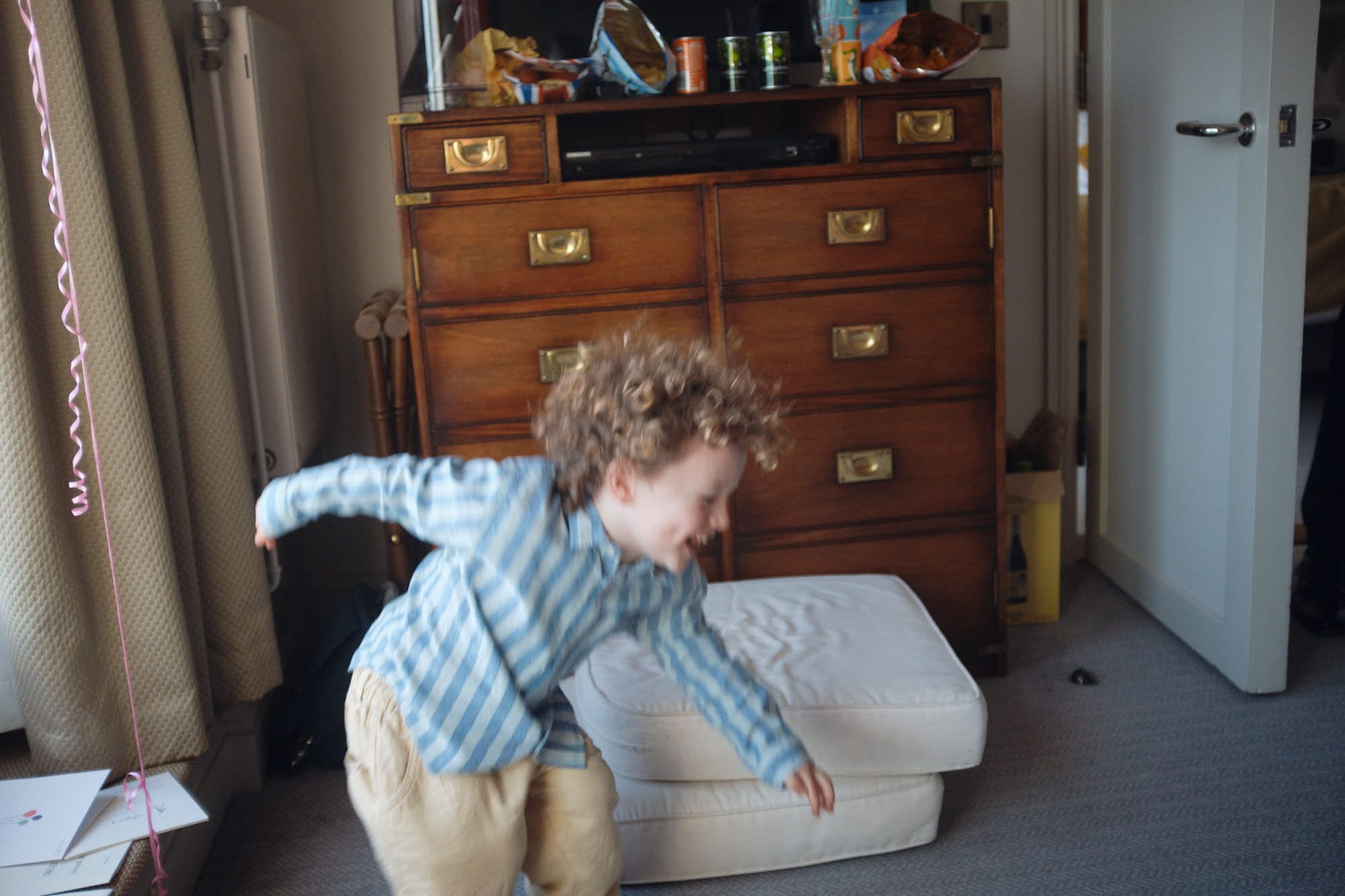 A young boy with curly hair and a blue and white striped shirt is running towards the camera, his arms outstretched and a joyful expression on his face. He is in a small, dimly.