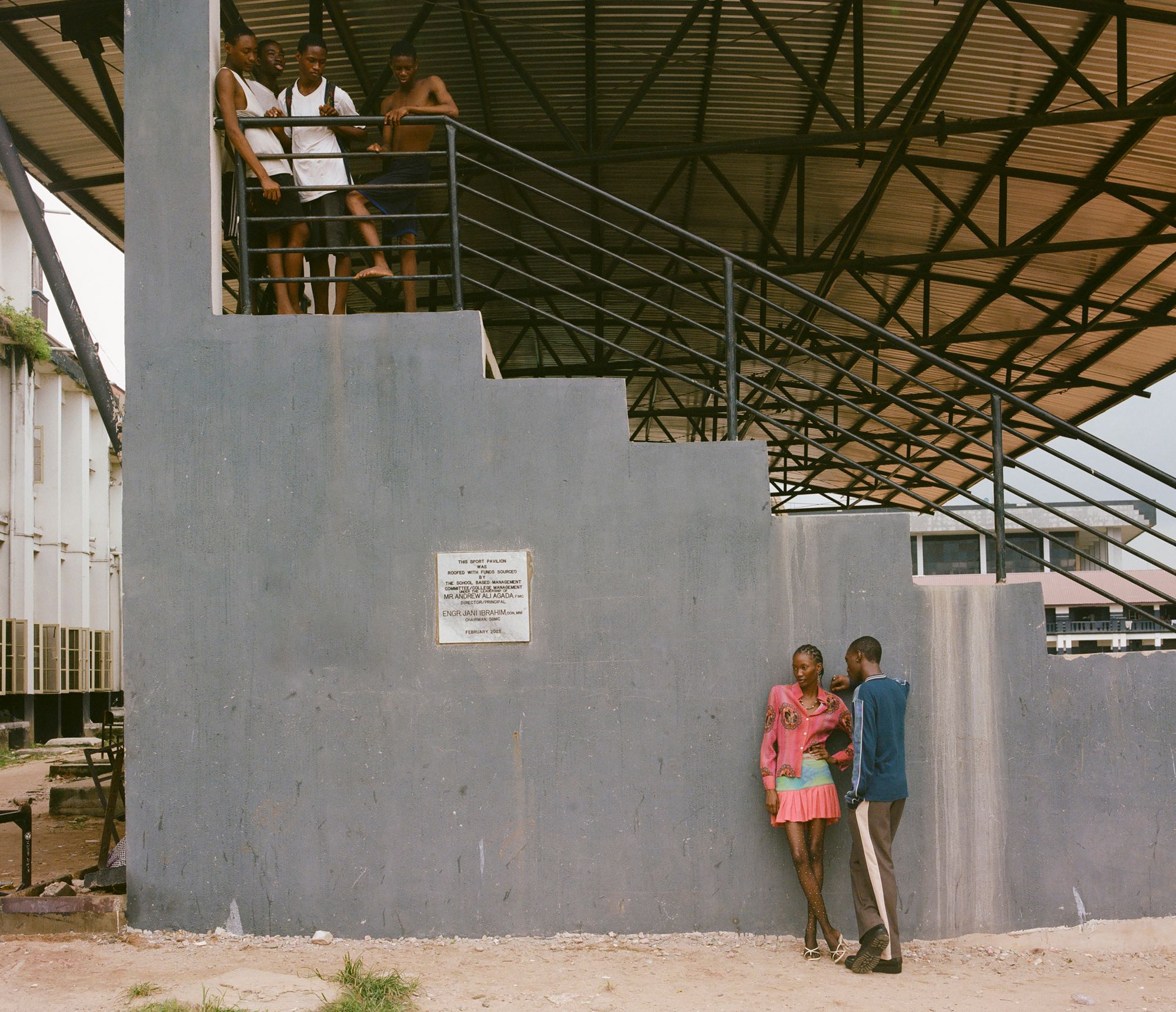 A group of young people stand on a metal staircase in an industrial building, with others in the background.