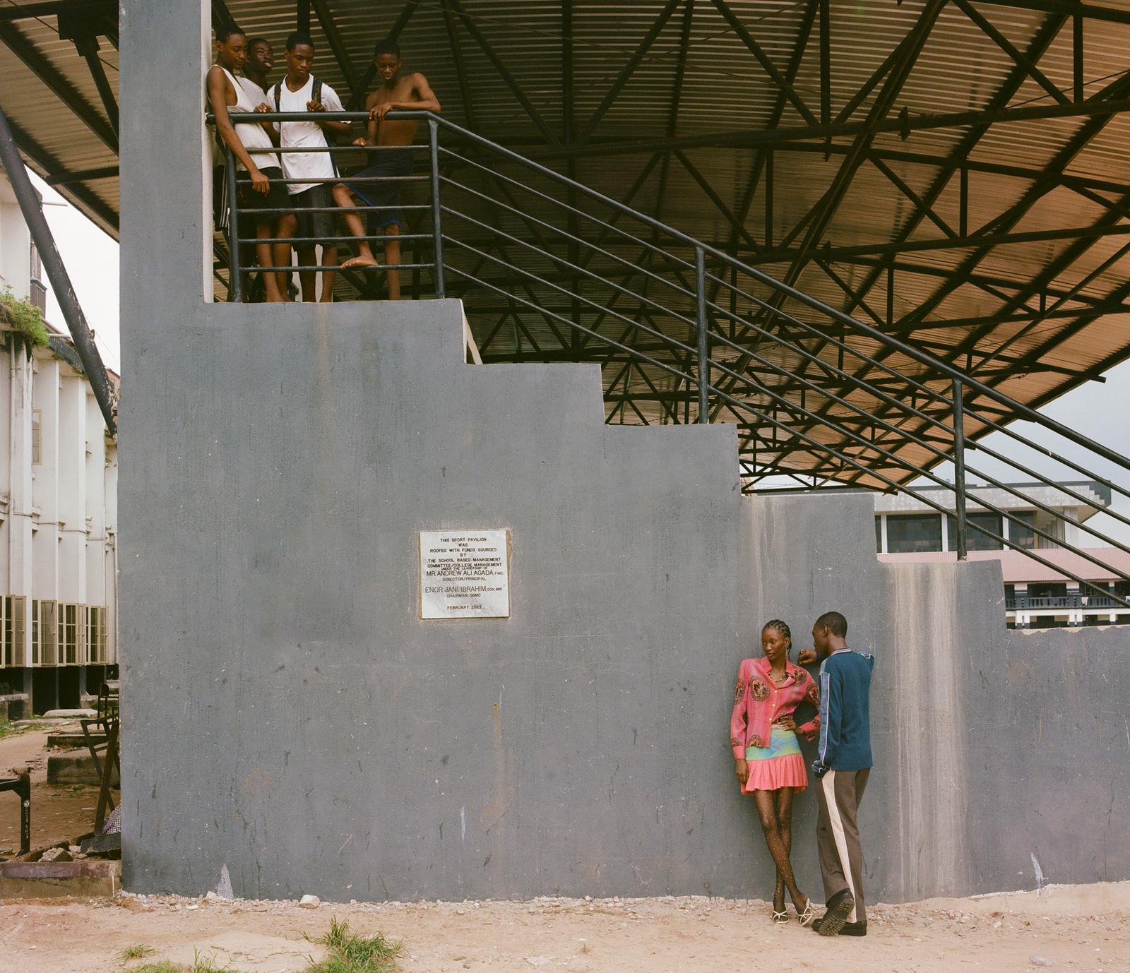 A group of young people stand on a metal staircase in an industrial building, with others in the background.