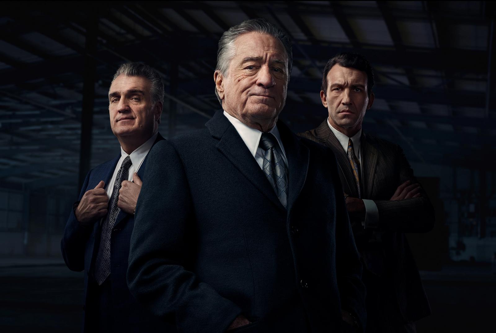Three men in suits, likely actors, posing in a dimly lit, industrial-style setting, with a background of metal beams and a brick wall.