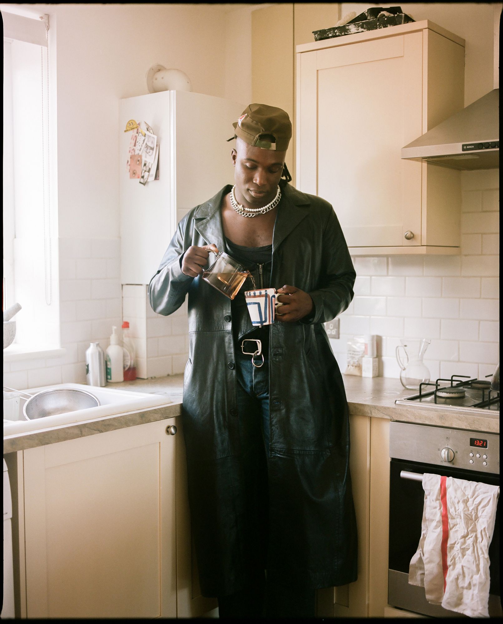 A person in a kitchen, wearing a black leather jacket, pouring tea from a teapot into a cup.