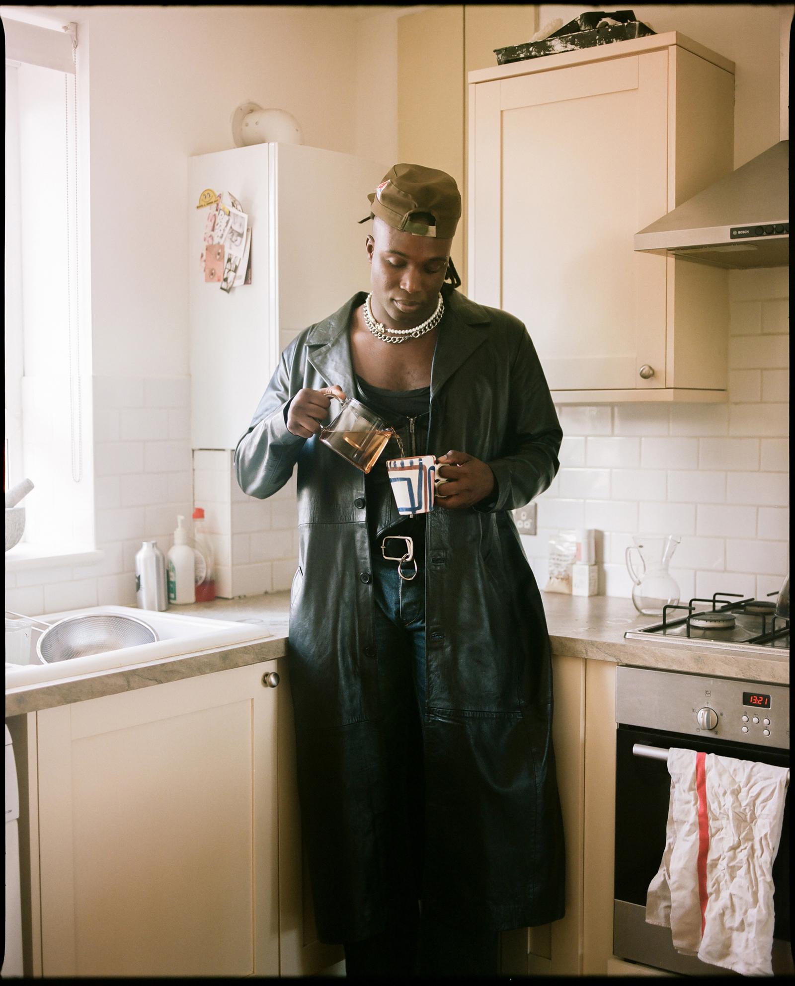 A person in a kitchen, wearing a black leather jacket, pouring tea from a teapot into a cup.