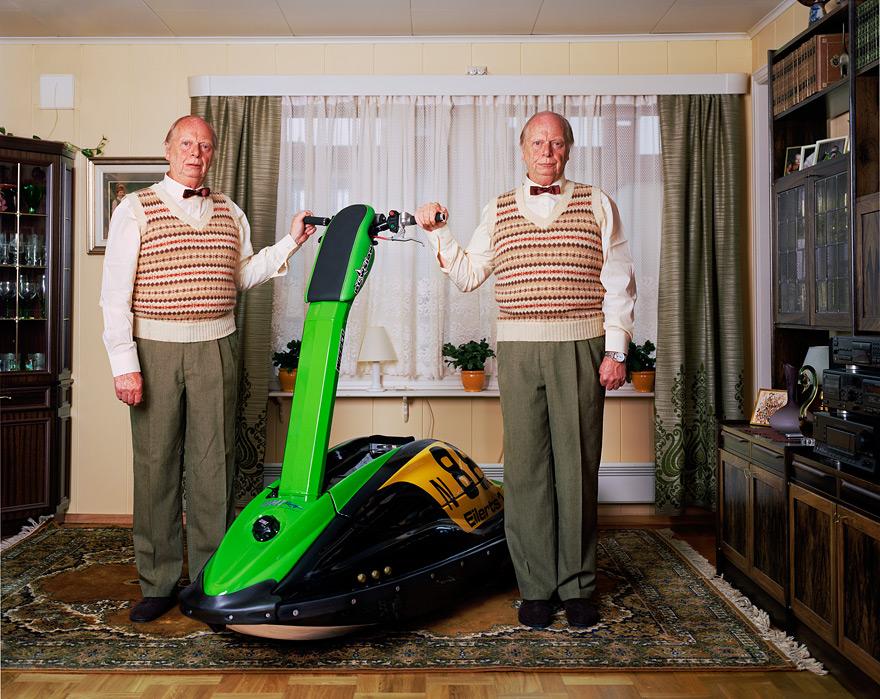 Two elderly men in identical outfits, one holding a green and black scooter, in a living room with a window and a TV in the background.